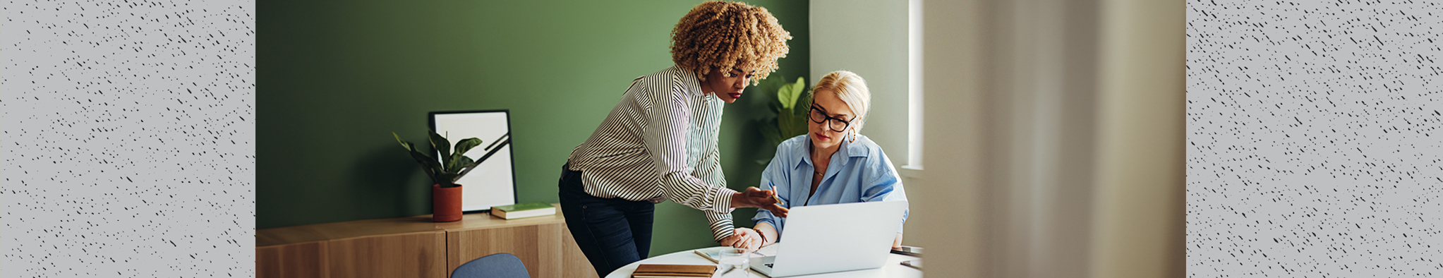 Two people speaking over a laptop, one recieving help from another in an office setting with a green wall background